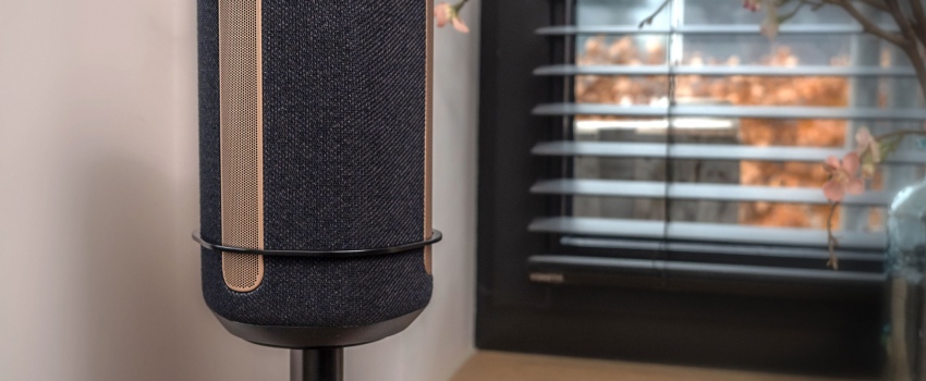Close-up of a black speaker on a Vebos stand with a bracket, placed in a modern interior near a window with blinds.